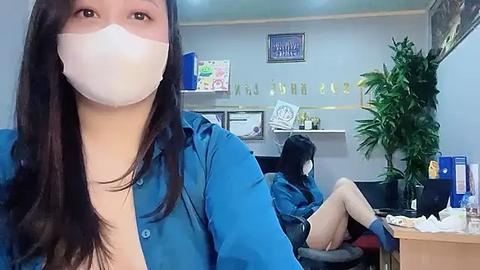 Media: Video of a woman with long dark hair in a blue shirt and mask sitting in a medical office. Another person is seated behind a desk, and plants and framed pictures adorn the walls.