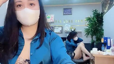 Media: Video of an Asian woman in a blue shirt, face mask, and glasses taking a selfie in an office with a potted plant, desk clutter, and a colleague seated.