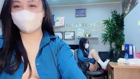 Media: A video of two women in an office. The foreground shows a woman with long black hair in a blue shirt, wearing a mask. The background features another woman in a black shirt, also wearing a mask, sitting at a desk.