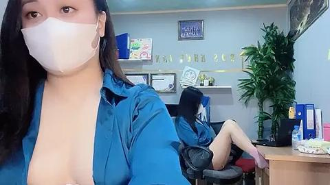 Media: Video of a woman with long dark hair, wearing a blue shirt and face mask, standing in a medical office with two other women seated at desks, plants, and certificates on the wall.