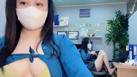 Media: Video of a woman with long black hair, wearing a blue shirt tied at the chest, a white face mask, and a yellow bra. Background shows two women, one in a similar outfit, seated in an office.