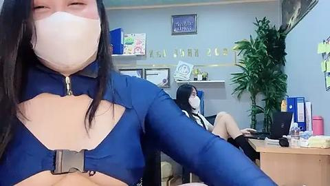 Media: Video of a woman in a blue, zipped-up, high-collared outfit with a white face mask, sitting in an office with two other women in similar attire. The background features a desk, potted plant, and office supplies.