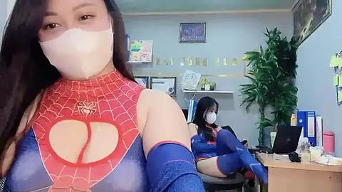 Media: Video of two women in Spider-Man costumes, one posing in front, the other seated at a desk in an office with potted plants, papers, and a computer.