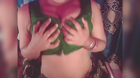 Media: A video of a woman with light skin, wearing a green top and a pink scarf, hands over her small breasts. She has a silver bracelet on her wrist. Background features a blue curtain with white floral patterns.