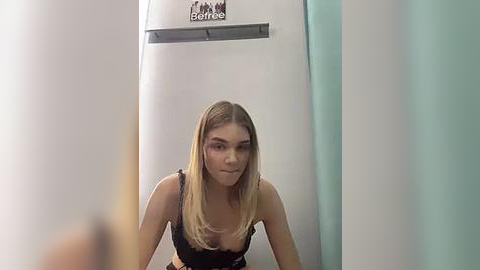 Media: Video of a young Caucasian woman with long blonde hair, wearing a black bra, standing in a bathroom with a beige Perbacco shampoo bottle on the wall.