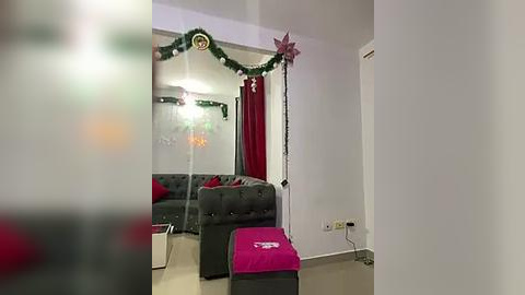 Media: Video of a minimalist living room decorated for Christmas with a dark gray sofa, a pink ottoman, green garlands, a large mirror, and festive red curtains.