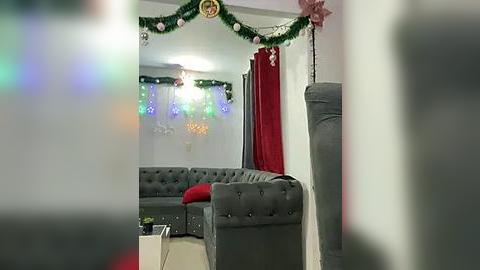 Media: Video of a cozy living room decorated for Christmas with a gray tufted sofa, festive garland, red curtains, and twinkling lights.