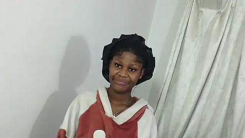 Media: Video of a young Black girl with short curly hair, wearing a black bonnet and a red and white hoodie, standing against a white wall and white curtain.