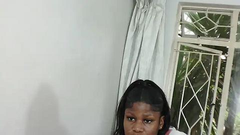 Media: A video of a young girl with braided hair, sitting against a white wall beside a window with a white curtain and diamond-patterned glass.