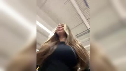 Media: A video captures a blonde woman with large breasts in a black top, standing in an industrial setting with a high ceiling and fluorescent lights. Her hair partially covers her face.