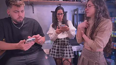 Media: Video of three young adults in a modern, well-lit kitchen. A man in a black t-shirt and chain necklace holds a phone, a woman in a white sweater and black plaid skirt looks puzzled, and a woman in a beige sweater and checkered skirt gestures.