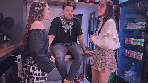 Media: Video: Three young adults in a dimly-lit convenience store. Two women in plaid skirts, one in a black off-shoulder top, the other in a white sweater. A man in a black t-shirt and gray jeans sits on a counter, wearing a scarf and glasses.