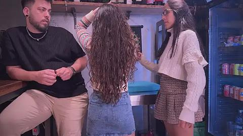 Media: Video: A tattooed man in a black shirt, a woman with long curly hair, and a woman in a white sweater and patterned skirt in a kitchen with colorful cabinets.