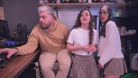 Media: Video: A bearded, gray-haired man in a tan hoodie and beige pants sits at a cluttered desk, with two teenage girls in school uniforms, one with curly hair and the other with glasses, standing beside him.