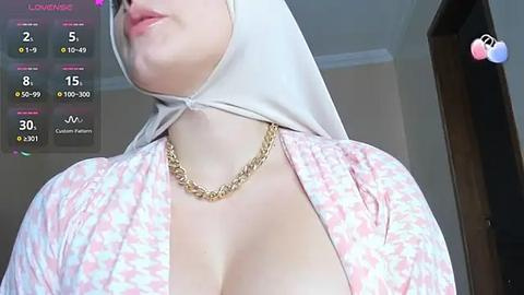 Media: A video of a fair-skinned woman with a large bust, wearing a white hijab and pink floral-patterned robe, adorned with a gold chain necklace. She stands indoors, with a virtual reality interface displaying her statistics in the background.