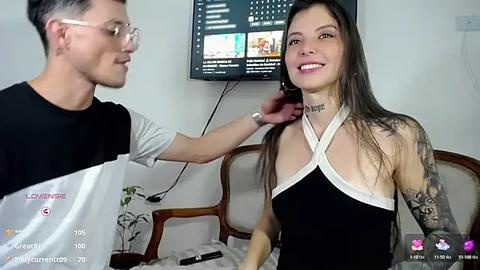 Media: Video of a young Asian woman with long dark hair, tattoos, and a black-and-white halter top, smiling while a young man with glasses adjusts her hair. Background includes a TV showing a game stream.