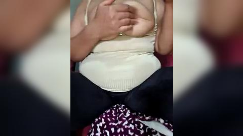 Media: Video of a pregnant woman in a beige tank top and black pants, sitting on a red-patterned blanket. Her hands are clasped over her belly.