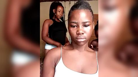 Media: Video of a young Black girl with short hair and light skin, wearing a white tank top, looking serious and contemplative. A darker-skinned girl with braided hair in a black tank top and white shorts stands behind her, adjusting her hair.