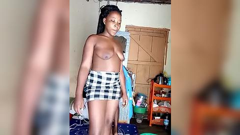 Media: Video of a topless, dark-skinned woman with medium-sized breasts, wearing a plaid skirt, standing in a cluttered, dimly lit room with a wooden door and shelves.