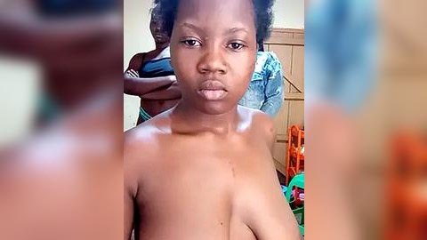 Media: Video of a young Black woman with dark skin and natural hair, topless, looking serious, indoors with blurred background.