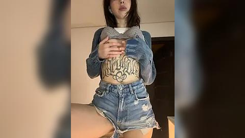 Media: Video of a young Asian woman with short black hair, wearing a grey cropped sweater and distressed denim shorts, revealing a tattoo across her midriff, taken indoors with blurred silhouettes in the background.