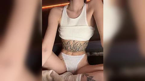 Media: Video of a slender, fair-skinned woman with short hair, wearing a white crop top and matching lace panties, sitting on a bed with a tattoo on her stomach, blurred faces in the background.