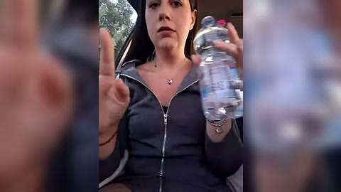 Media: A video of a young woman with short dark hair and fair skin, wearing a dark zip-up hoodie, sitting in a car. She holds a water bottle and gestures with her hands, set against a blurry background.