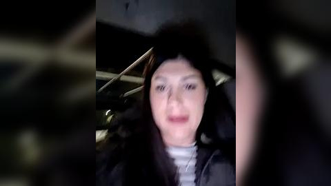 Media: A blurred video of a woman with long, dark hair, wearing a grey top, sitting in a dimly lit, cluttered room with a wooden chair and metal objects in the background.