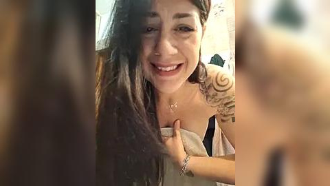 Media: Video of a young woman with long dark hair and a tattoo on her shoulder, smiling, holding a towel, in a dimly lit room.