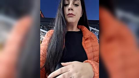 Media: Video of a woman with long black hair, wearing an orange puffer jacket and black top, holding a black object, in front of a blurred, dark background with a blue sky visible.