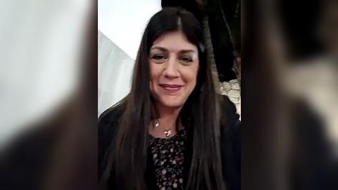 Media: Video of a smiling Latina woman with long, dark hair, wearing a black floral-patterned blouse, and a black blazer. The blurred background shows a white wall and a plant.