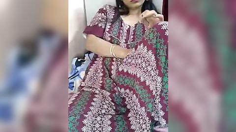 Media: Video of an Indian woman in a maroon and white patterned sari, seated indoors with a blurred background.