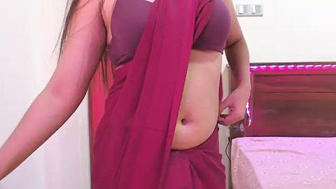 Media: Video of a woman with long, dark hair, wearing a purple sari, adjusting the fabric around her midsection in a bedroom with a bed, pink sheets, and a wooden headboard.