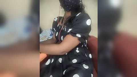 Media: Video of a dark-skinned woman with long braids, wearing a black dress with white polka dots and a headscarf, sitting at a table, blurred background.