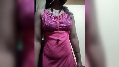 Media: Video of a woman wearing a pink sleeveless dress with lace detailing, black headphones, and a necklace, taken indoors with blurred background.