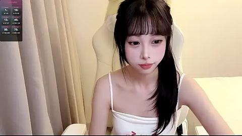 Media: Video of a young East Asian woman with long black hair, wearing a white spaghetti-strap top, sitting in a beige chair against a cream-colored wall.