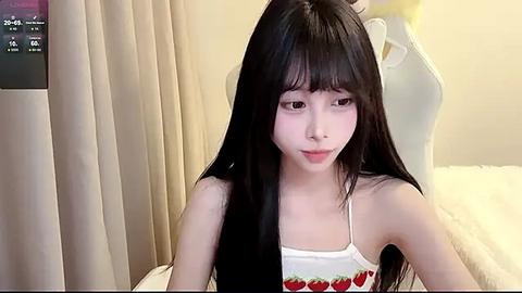 Media: A video of a young East Asian woman with long, straight black hair and fair skin, wearing a white tank top with strawberry print, sitting indoors, looking contemplative.