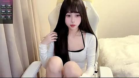 Media: Video of an Asian woman with long black hair, sitting in a white gaming chair, wearing a white long-sleeve top, black bra, and beige pants.