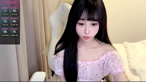 Media: A video of an East Asian woman with long black hair, wearing a pink off-shoulder top, sitting on a white chair in a room with beige curtains and a mannequin.