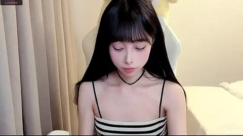 Media: A video of a young Asian woman with long black hair, wearing a black and white striped tank top, sitting on a white gaming chair in a minimalist bedroom with beige curtains.