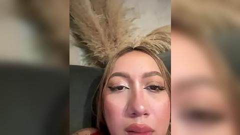 Media: A close-up video of a woman with fair skin, wearing dramatic feathered headwear and heavy makeup, including exaggerated eyeliner, on a dark background.