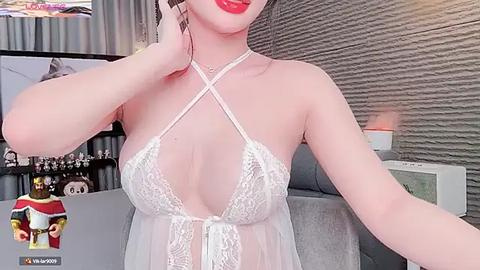 Media: Video of an East Asian woman with fair skin, wearing a sheer white lace lingerie top with crisscross straps, red lipstick, and holding a phone. Background shows a modern living room with grey decor, shelves, and a window.