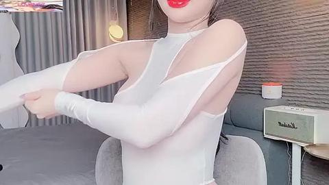 Media: Video of an East Asian woman with fair skin, wearing a white, long-sleeved, sheer bodysuit, red lipstick, and dark hair, adjusting her sleeve in a modern living room with gray carpet, gray couch, and textured gray wall.