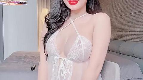 Media: Video of an Asian woman with long black hair, wearing a sheer white lace lingerie top, red lipstick, standing in a modern bedroom with a grey bed and textured brown wall.