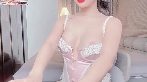 Media: Video of an Asian woman with fair skin, wearing a revealing pink satin corset with white lace trim, sitting on a grey bed in a modern bedroom.