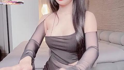 Media: Video of a slender, light-skinned woman with long black hair, wearing a sheer black sleeveless top and matching sheer sleeves, standing in a modern, softly lit bedroom.