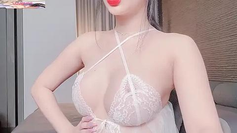 Media: A video of an Asian woman with fair skin, wearing a lacy, white, halterneck bra revealing ample cleavage, red lipstick, and long, straight hair. She stands indoors near beige curtains and a bed.