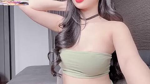 Media: Video of a fair-skinned Asian woman with long, wavy black hair, wearing a strapless olive-green bandeau top, black choker, and red lipstick, posing with one arm raised. Background shows a gray sofa and textured wall.