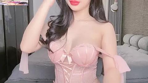 Media: Video of a fair-skinned woman with long, dark hair in a pink corset with lace, posing indoors. She wears red lipstick and stands near a grey bed with a tufted headboard.