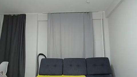 Media: Video of a minimalist room with a black, yellow, and gray sofa against a white wall, featuring a large gray curtain and a black curtain to the left.